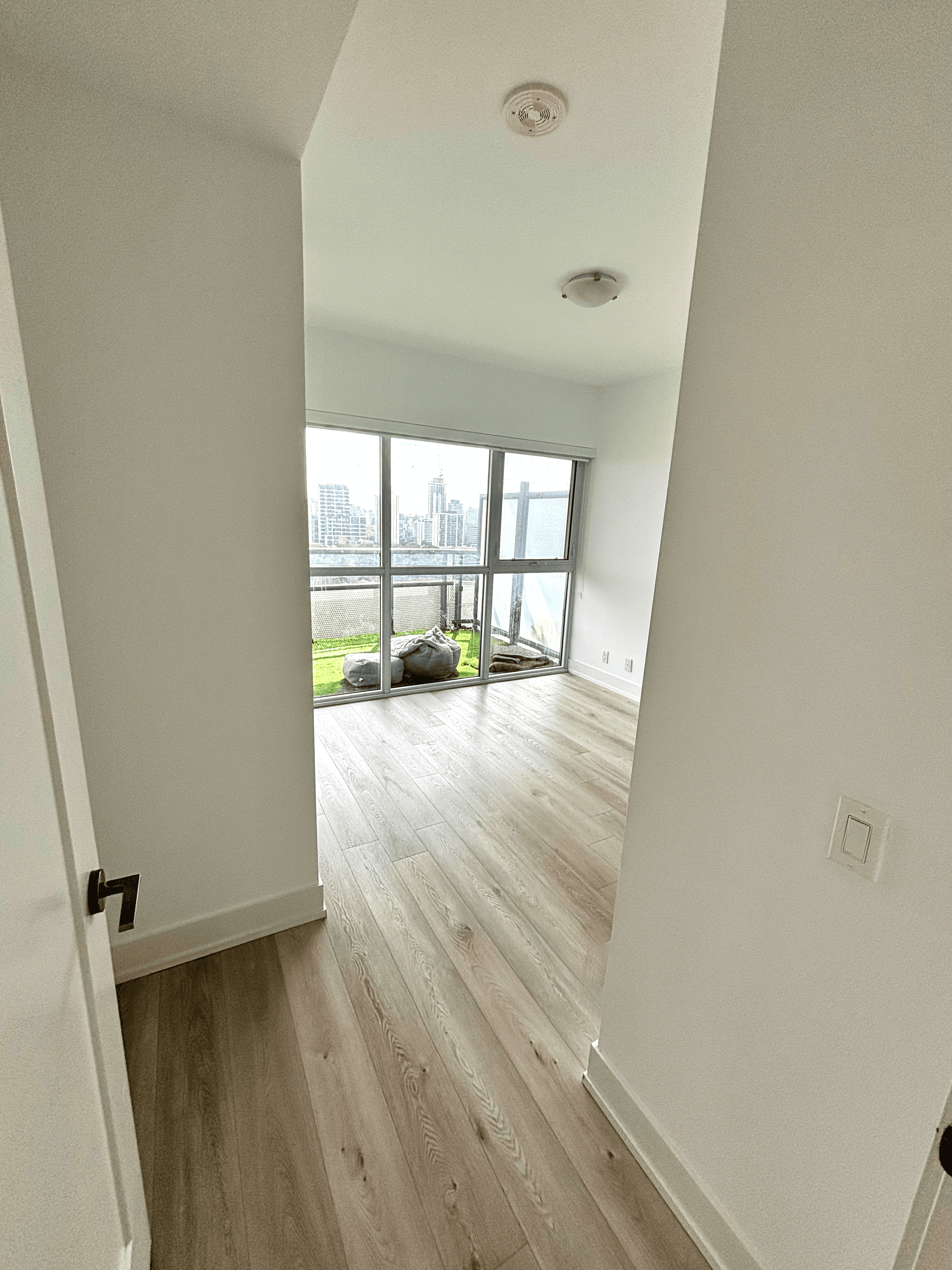 Bright empty room with light wood floors and large windows overlooking a city skyline.