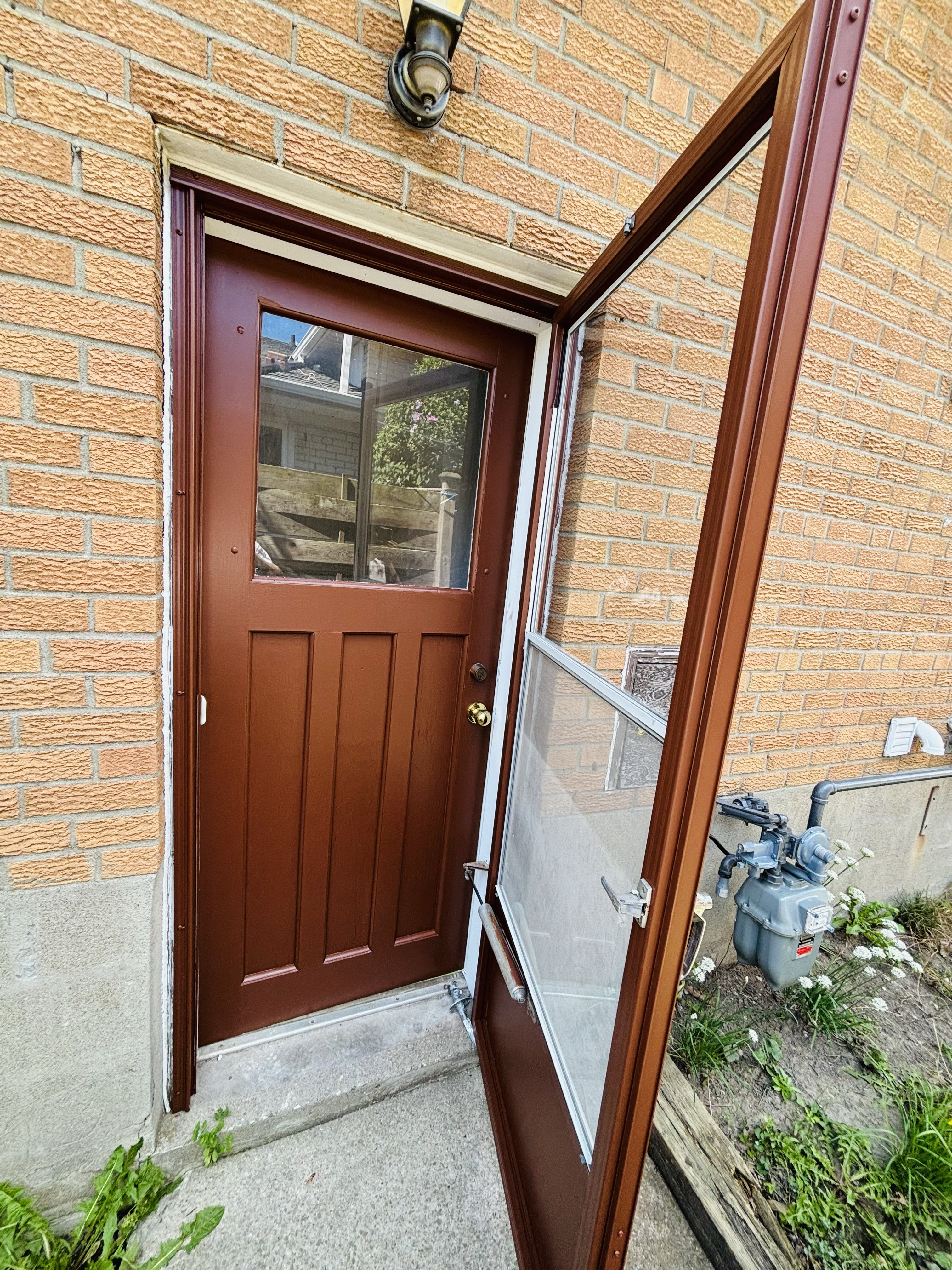 Brown exterior door with window and open storm door against a tan brick wall.