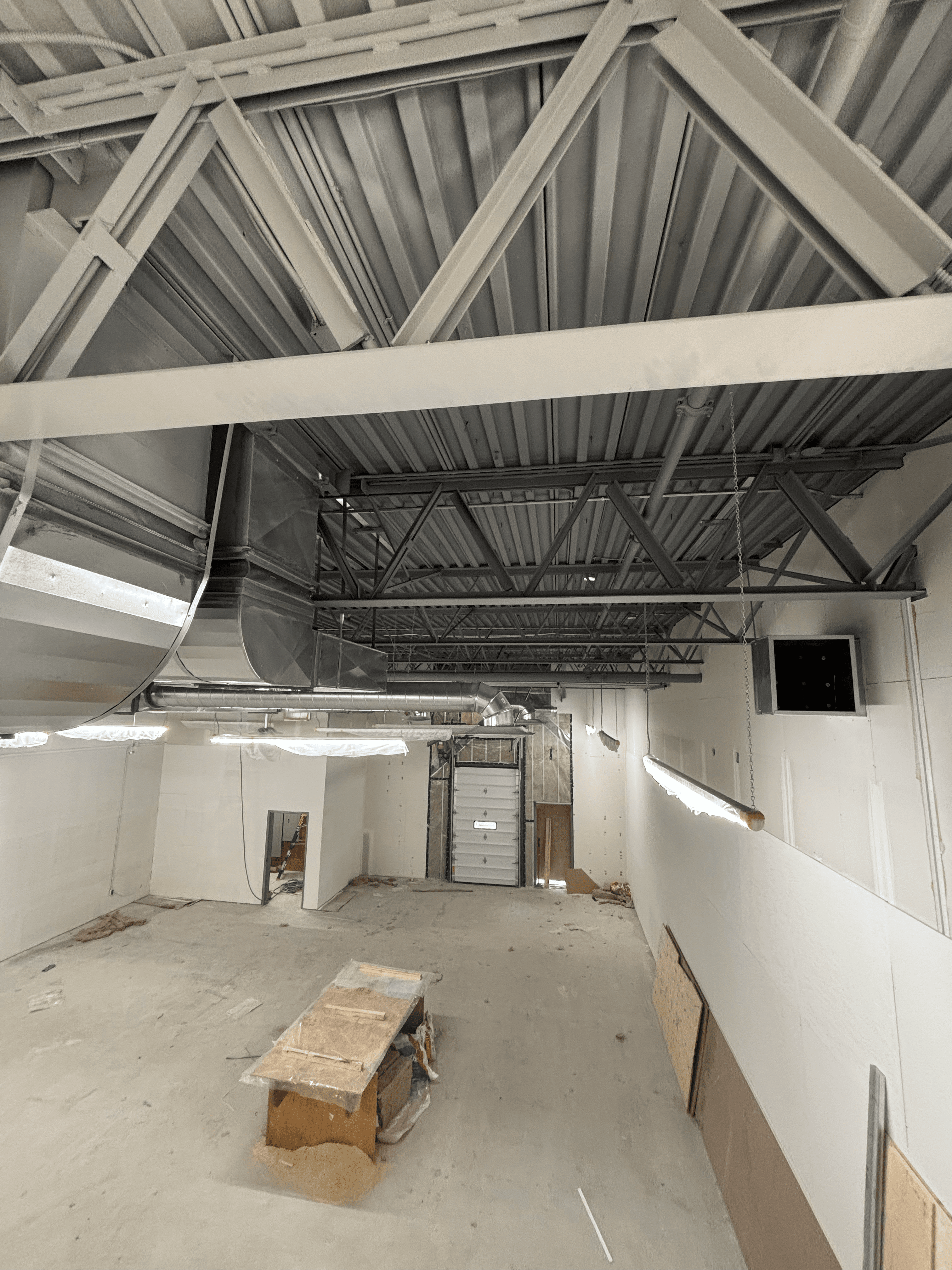 Industrial warehouse interior under construction with exposed ceiling trusses, metal ductwork, and a workbench.