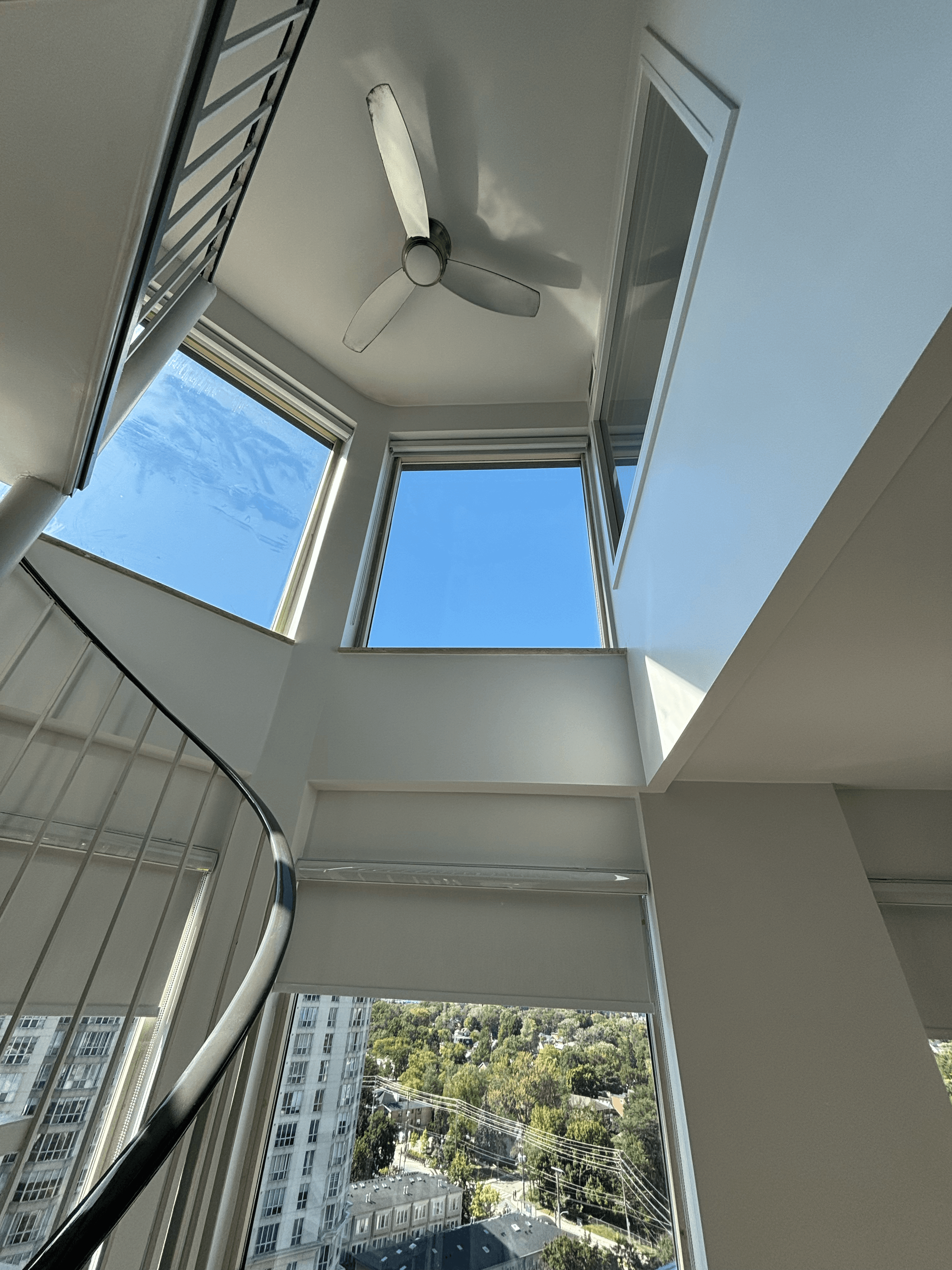 Spiral staircase and ceiling fan in a modern white room with windows overlooking a city.