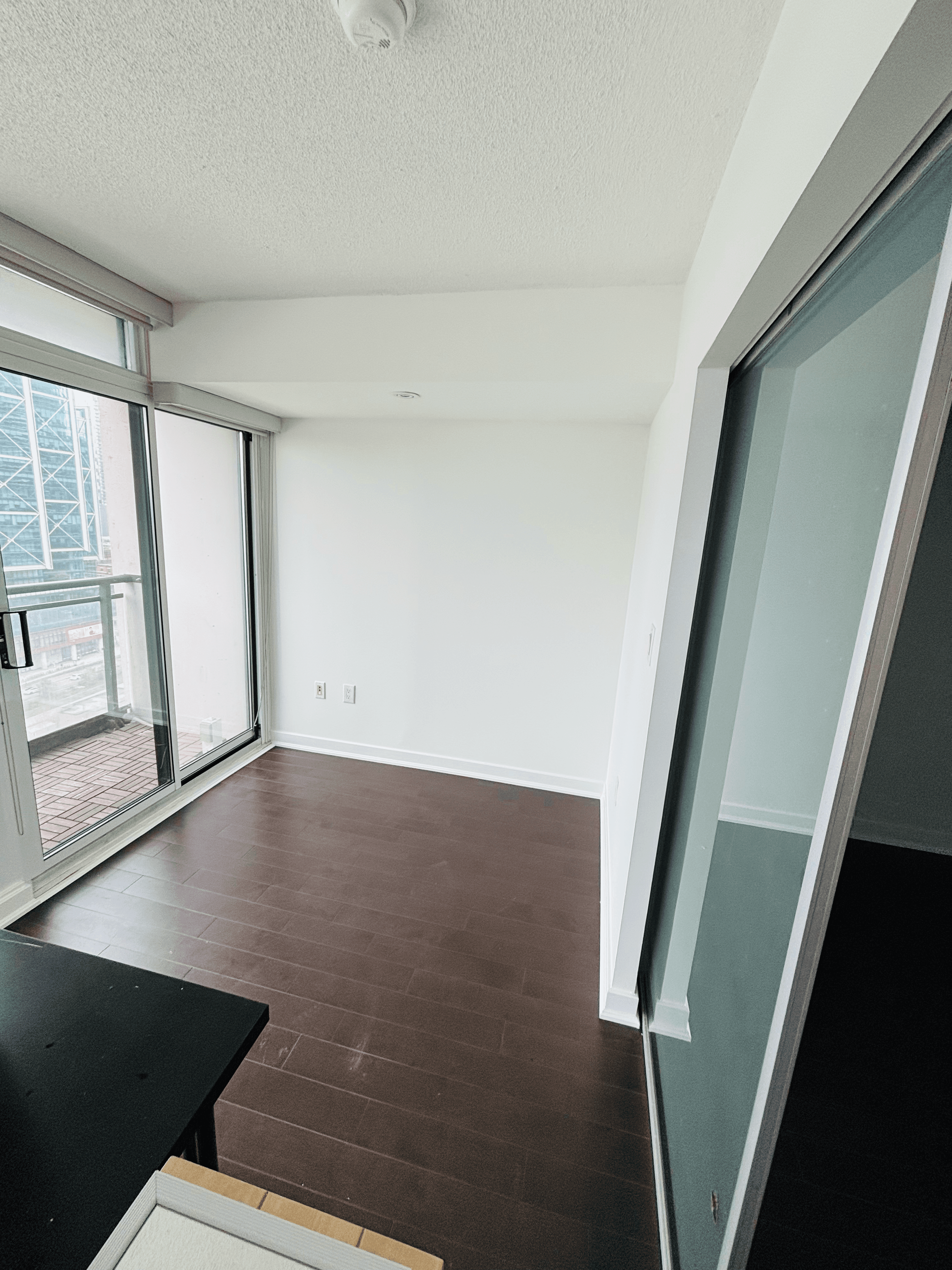 Empty apartment room with dark wood floors, white walls, and large windows overlooking a balcony.