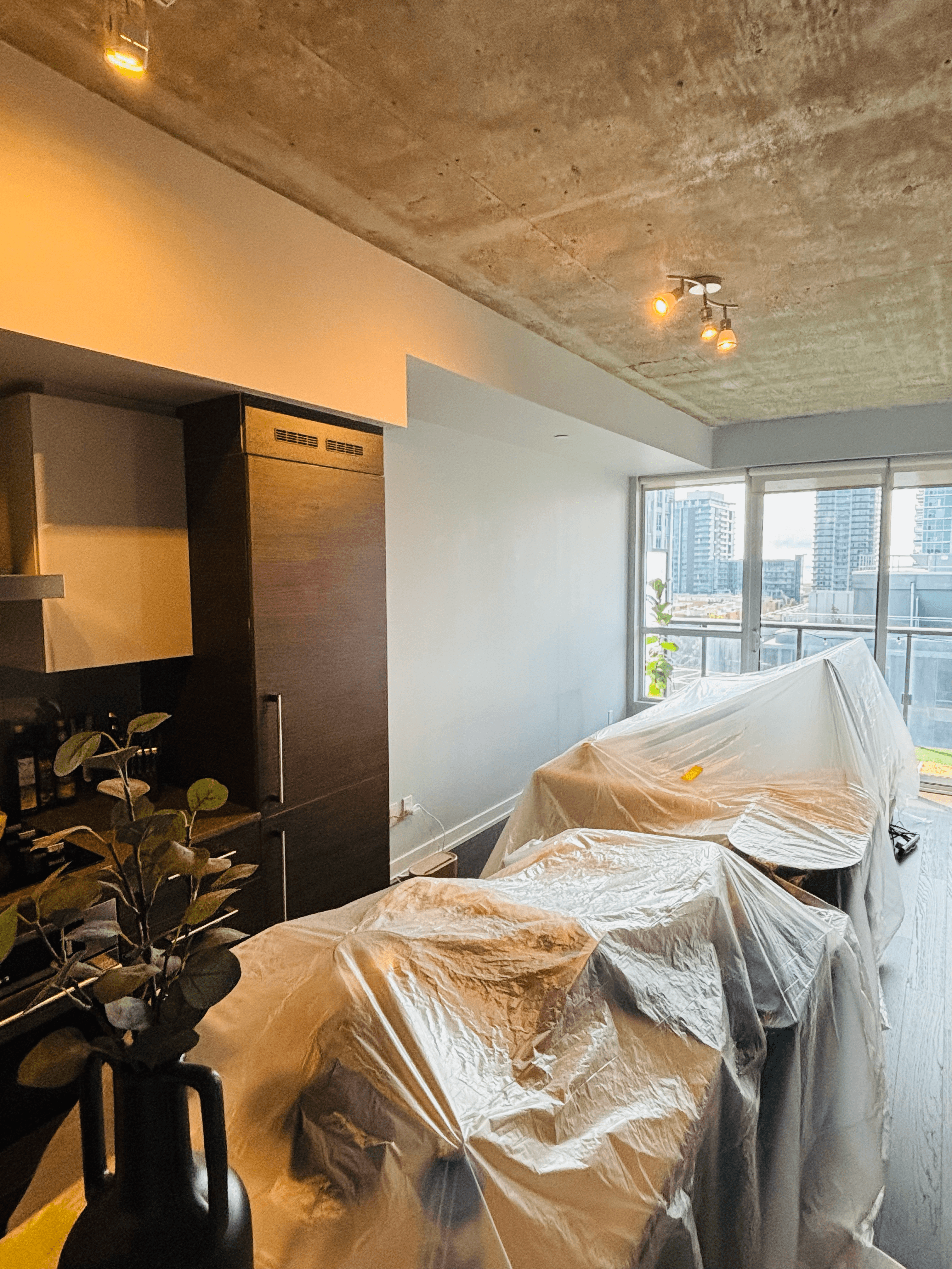 Furniture draped in plastic drop cloths inside a modern apartment overlooking a city skyline.
