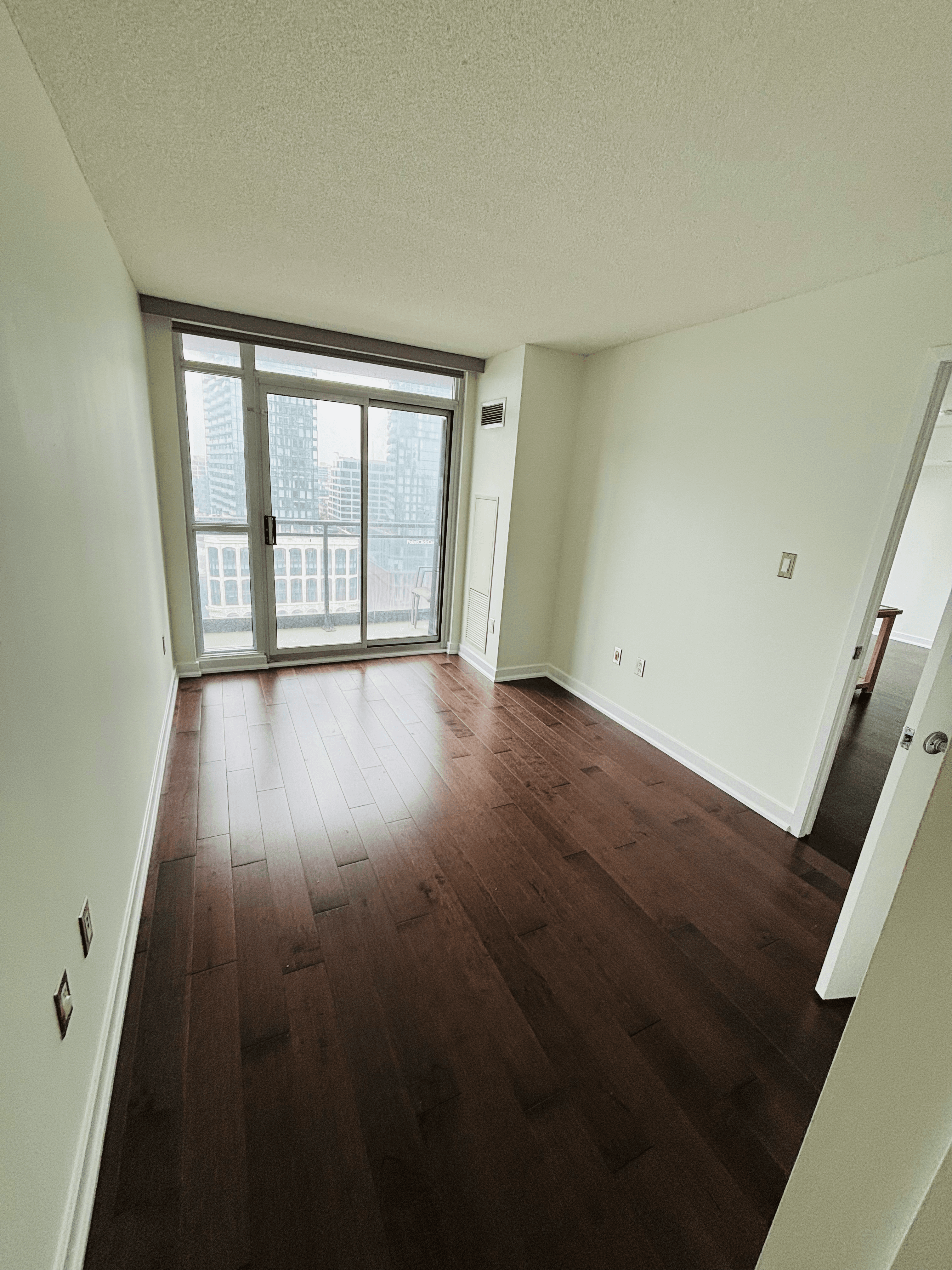 Empty apartment room with dark hardwood floors and sliding glass doors overlooking city buildings.