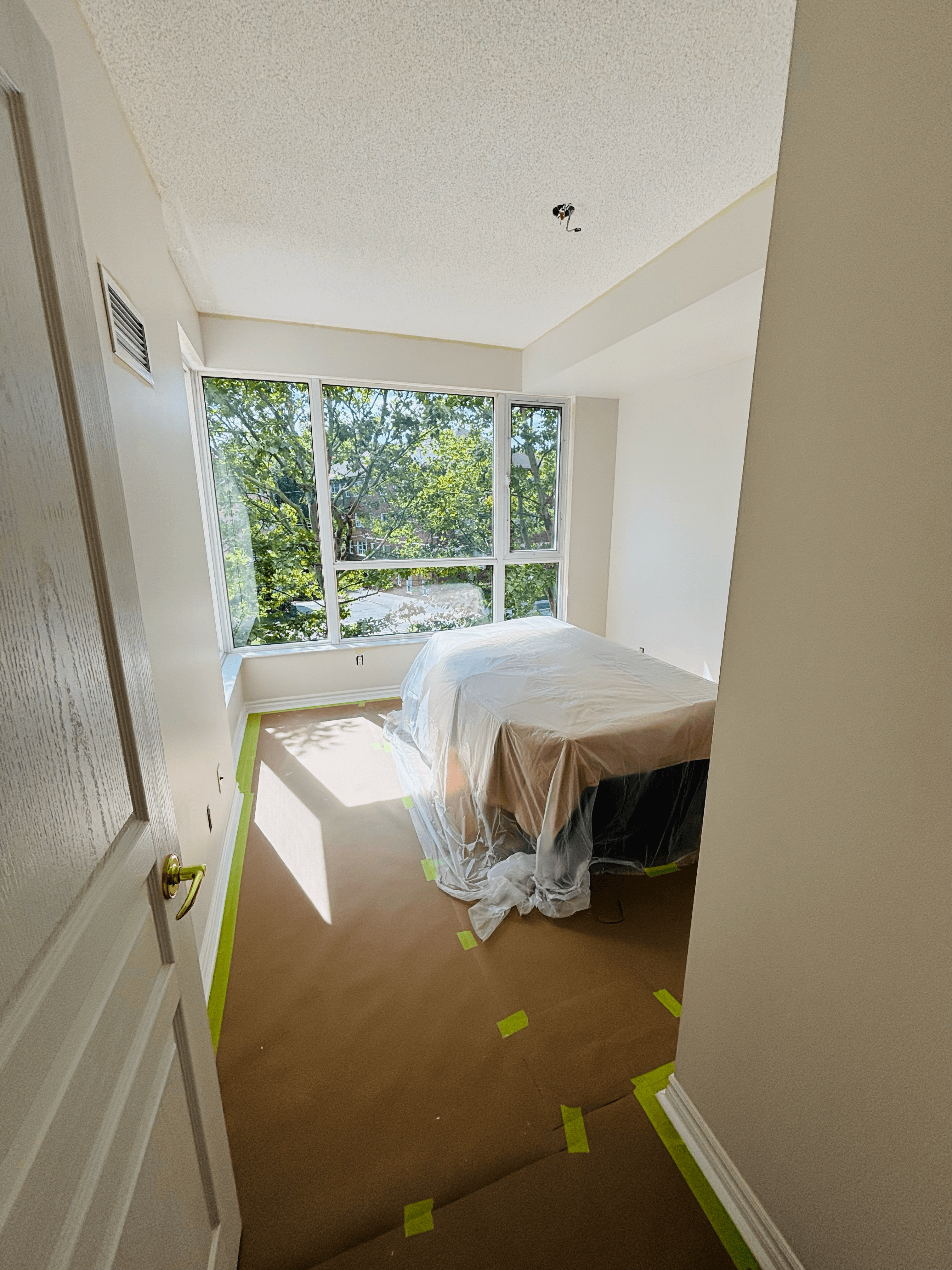 Bedroom under renovation with plastic-covered bed, paper-protected floors, and large windows overlooking green trees.