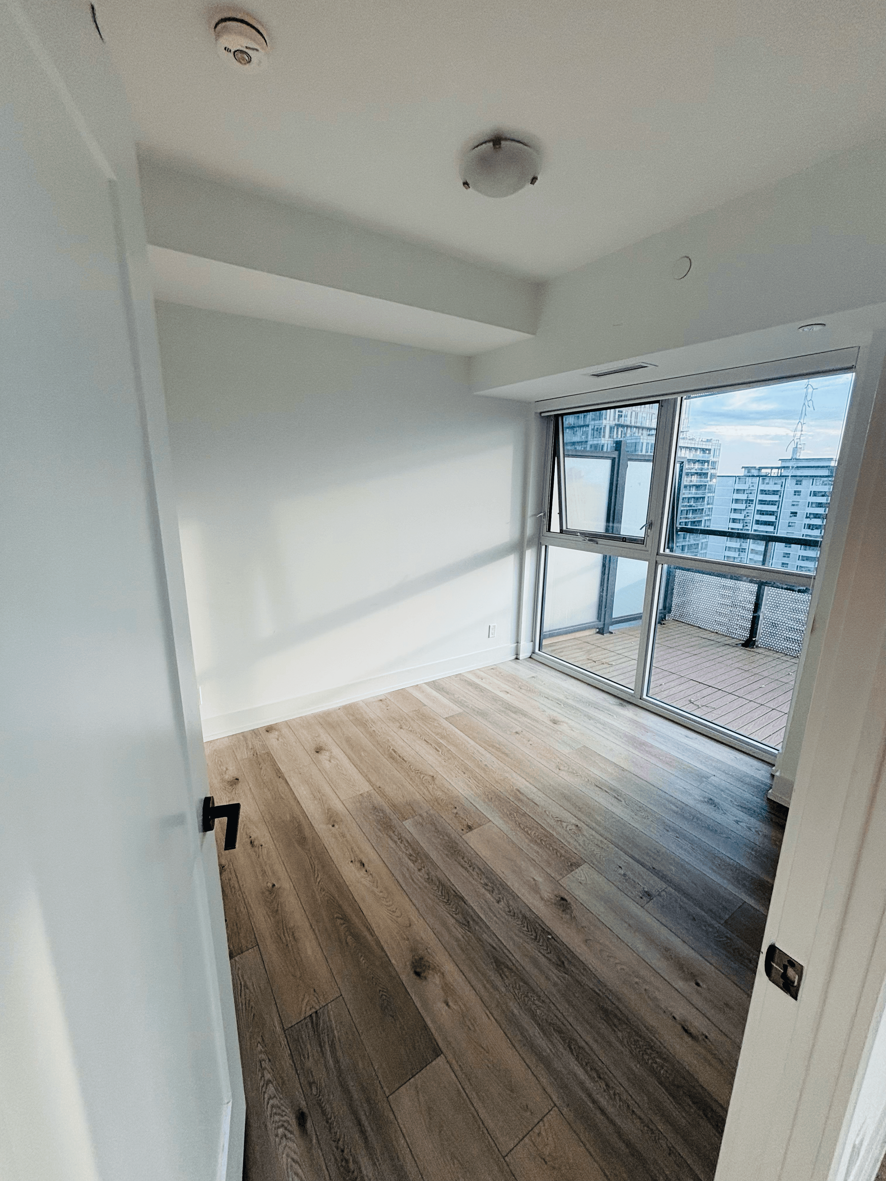 Modern empty room with light wood flooring and glass doors leading to a city balcony.